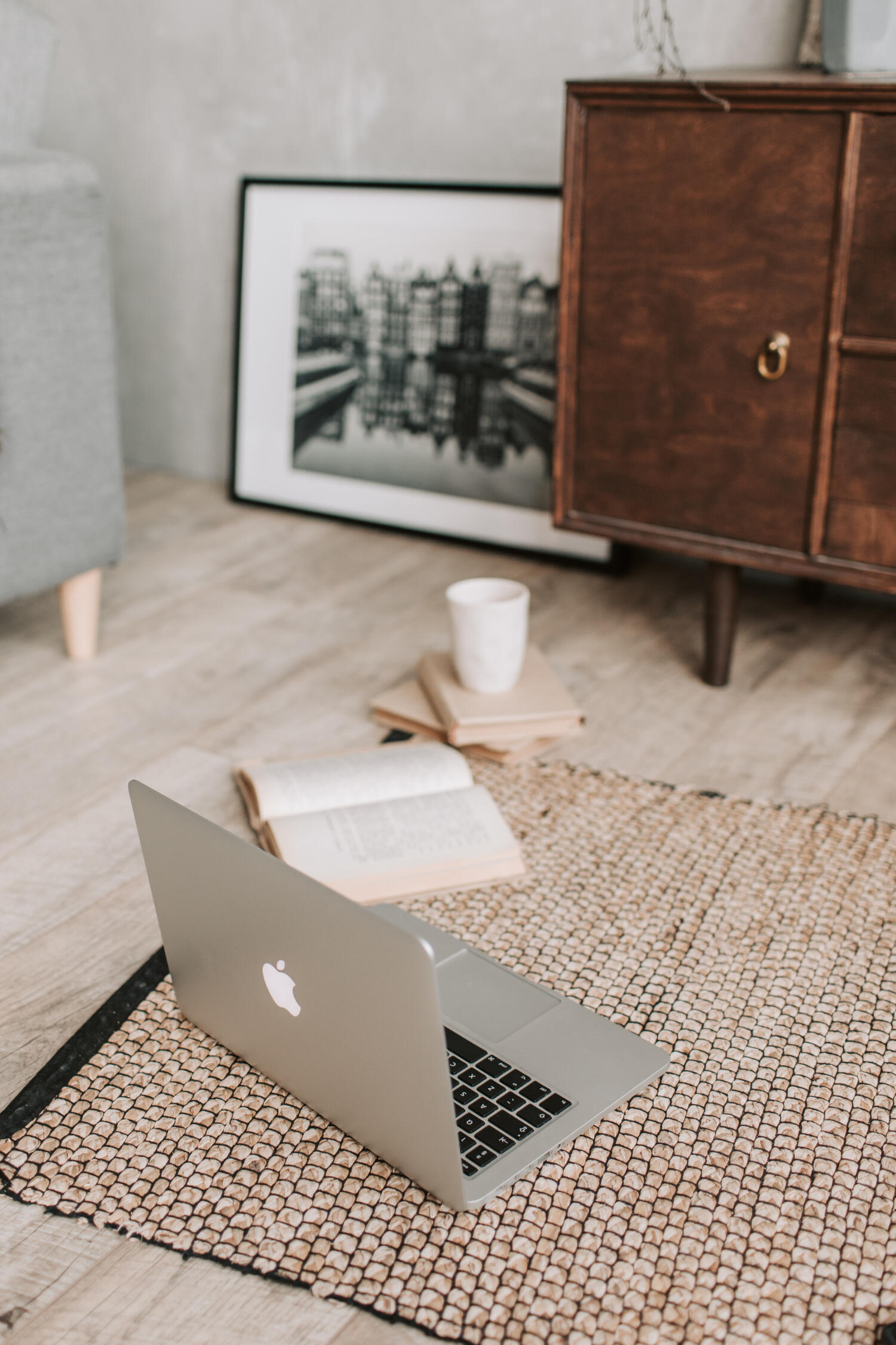 A laptop, old book, and mug on a light wood hardwood floor with a black and white print leaning against the wall between a light grey couch and dark brown antique sideboard.